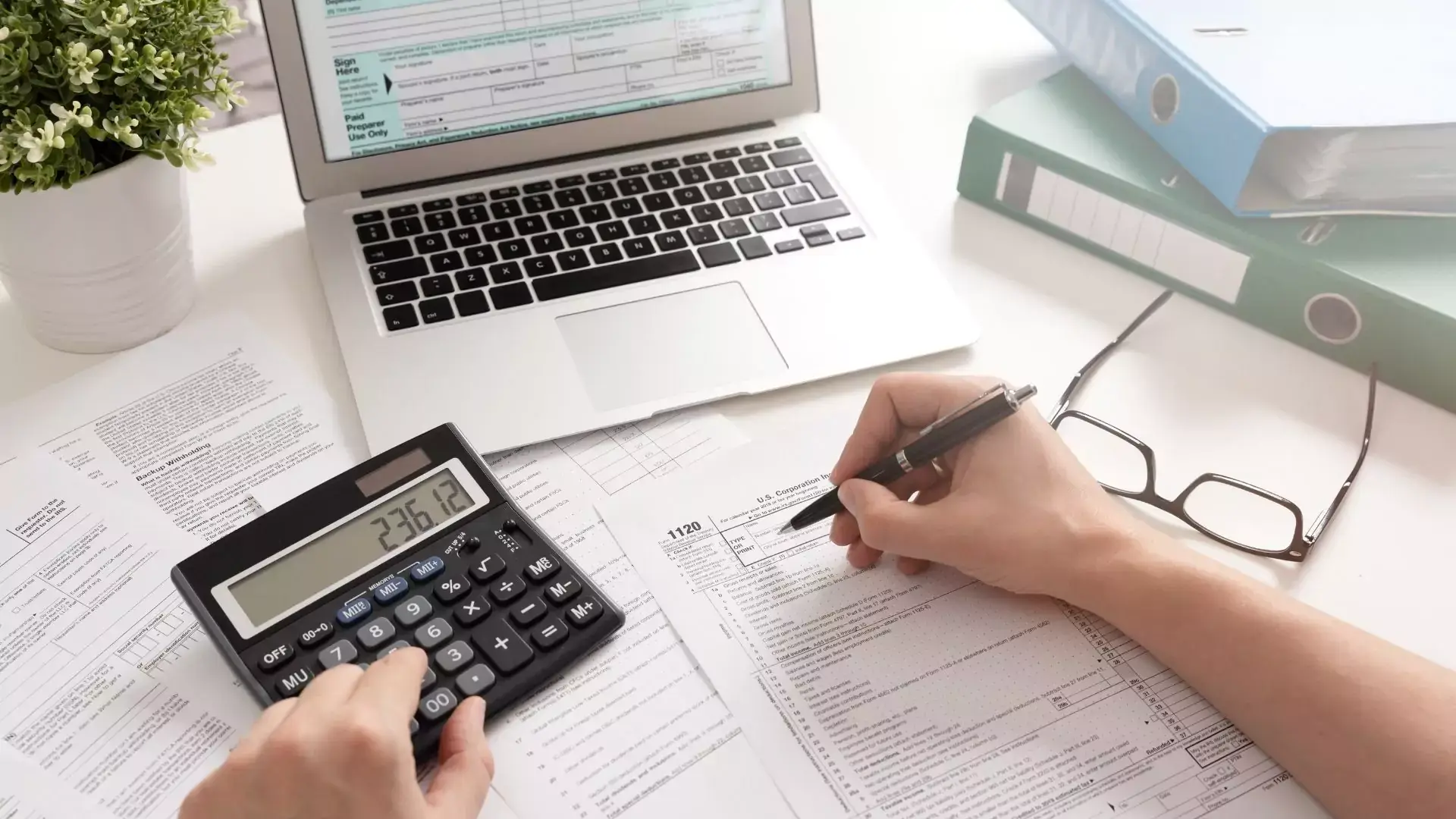 A laptop and calculator on a desk and a person writing a report