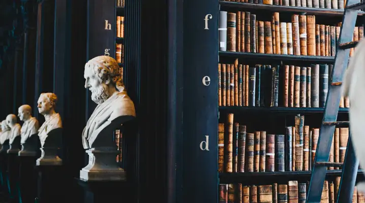 A row of stone sculptures, full bookshelves, and a wooden ladder in a library